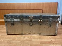 Closed aluminium steamer trunk showing front face with multiple rivets, metal latches, lock, and ring handle. Signs of wear and dents are visible.