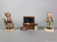 Three Goebel porcelain figurines with small wooden chalkboard display and school-themed miniature accessories, all on a flat surface with grey background.