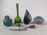 Front view of all five items including the green glass vase, black pottery pitcher, blue-brown lidded pottery jar, metal and teak cheese cutter, and metallic kinetic paperweight.