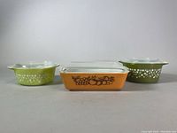 Front view of three covered Pyrex dishes showing colors and patterns: chartreuse green with white daisies, orange with fruit decal, light green with white daisies