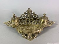 Front angle view of the brass Gothic Revival inkwell dip stand showing the two lidded inkwells, ornate filigree body, and detailed devil imp face decoration on the backplate.