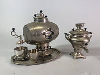 Photo showing barrel-shaped mini samovar on attached tray with two metal cups and saucers and smaller urn-style samovar, illustrating the complete set.