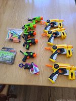 Photo showing six yellow and black Nerf pistols, four smaller green/orange/black mini blasters, a clear bag of various foam darts, and a boxed pack of Zombie Strike darts arranged on a wooden table.