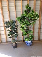 Two artificial trees with different foliage types and pots shown against wooden wall