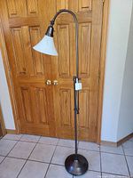 Full view of the tall adjustable OttLite floor lamp with bronze finish and white bell-shaped lampshade, standing on tile floor in front of wooden closet doors.