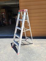 5-foot aluminum step ladder shown standing on pavement in bright sunlight with an open garage in the background. The step ladder has red caps on the feet and a red top platform.