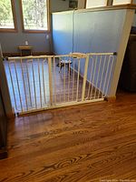 White metal baby/pet safety gate installed in a hardwood floor opening with wooden corner trims.