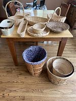 Nine assorted baskets on wooden table and floor including handled wicker, oval and rectangular shapes, plastic basket, and wooden bucket basket with liners.