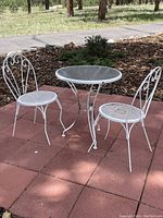 White metal bistro table with glass top and 2 matching chairs on outdoor red tile surface. Trees and greenery in background. Table and chairs show some rust but sturdy condition.
