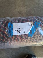 Rolled braided area rug wrapped in plastic with visible handwritten label showing dimensions 102" x 82" and sale price.
