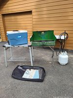 Photo showing overall camping setup with Coleman 3-burner stove, propane bottle and hoses, wheeled cooler, folding aluminum table, and black carrying case on asphalt surface outside building.