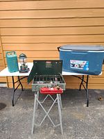 Photo showing Coleman 2-burner stove on gray metal folding stand, green Coleman gas lantern on white table, large blue Coleman Extreme cooler on white table, and a green hard plastic carrying case on the left side.