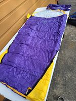 Purple and yellow lightweight youth sleeping bag laid open on a table outdoors, showing zipper and material.