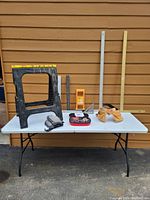 Photo of the saw horse, squares, ruler, tool belt, knee pads, and some measuring tools arranged on a white folding table against brown siding.