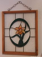 Framed stained glass flower window with orange flower and green leaves inside a wooden frame and chain hanging.