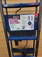 Photo showing HaulMaster appliance hand truck label with 600 lb. capacity and usage instructions attached to blue metal frame with black straps.