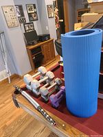 Photo shows four hex dumbbells of weights 10, 15, and 20 pounds, a pair of small purple dumbbells, and part of a blue rolled yoga mat, along with a spring bar with wooden handles on a wooden floor near a pool table.