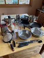 Overview of kitchenware items arranged on wooden table, presence of large pot, graters, strainers, skillets, measuring cup, and utensils