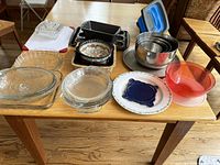Photo showing a collection of kitchenware including glass baking dishes, metal baking pans, stainless steel mixing bowls, a collapsible sieve, and various plates and bowls arranged on a table