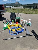 Photo showing various lawn care tools including spreaders, sprayers, hoses, fertilizers, and other miscellaneous items arranged on pavement outdoors with a mountain and fence in background.