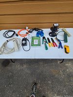 Photo of the entire lot arranged on a white outdoor table showing extension cords, timers, power strips, wire spools, single gang boxes, stud finder, and hand tools.