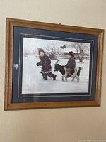Photo showing a framed picture hung on a wall, depicting two children dressed in winter clothing with a black and white goat in a snowy farm background.