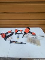 Photo showing three orange pneumatic nailers of different sizes, a clear plastic box with nails, a pack of nails, and a black pneumatic tool gauge on a white table against a brown wooden wall.