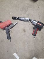 Three pneumatic tools laid on a concrete floor; from left to right: sand blaster tool with red container, silver 3/8 inch air ratchet wrench, black and red Craftsman 1/2 inch impact wrench.