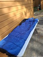 Full length view of the blue and black mummy sleeping bag laid out flat on a white surface outdoors against a tan wooden wall.
