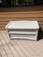 White plastic Suncast deck box on a wooden deck. The box has a flat lid, two side handles, and a panel design on the body. Used condition visible.