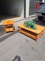Two white cherry wood tables; coffee table with two front drawers and end table with one drawer and open shelf, both placed outdoors on asphalt near a metal building.