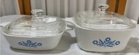 Two white Corning Ware casserole dishes with blue cornflower pattern and clear Pyrex lids. Both dishes have single handles, shown from front.