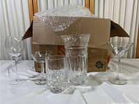 Full view of crystal and cut glass items arranged in front of a cardboard box with bubble wrap on top. Items include a large cut glass vase, tall stemmed glasses, and shorter cut glass pieces.