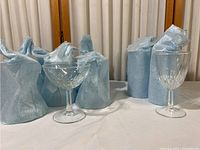 Two crystal glasses displayed with six others wrapped in blue protective paper. One coupe glass on the left and taller stem glass on the right.