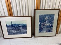 Two framed photo prints leaned against a wall showing European scenes, one horizontal and one vertical orientation.
