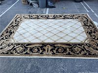 Full top-down view showing the entire rectangular rug laid out on pavement. Displays ornate gold and cream design bordered in black with floral and scroll patterns.