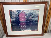 Framed photographic print showing a red house by a river with reflection in water, medium brown decorative wooden frame.