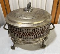 Front angle view of the vintage metal serving dish holder with lid showing the decorative cutwork pattern, side handles, and claw feet.