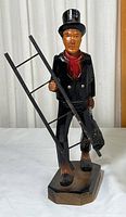 Front view of the wooden chimney sweep figurine holding a ladder and broom, showing painted black coat, red scarf, and black top hat.