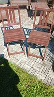 Photo of two foldable wooden chairs on tiled outdoor surface with grass edge visible, showing the front and side views.