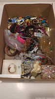 Cardboard box filled with assorted costume jewellery and crafting items, including bracelets, beaded necklaces, brooches, pins, and plastic hair combs.