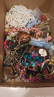 Box containing mixed costume jewellery and crafting materials in various bead colors and materials including white, pink, black, turquoise, wood, and plastic.