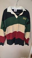 Front view of vintage rugby shirt with black, green, cream, and red horizontal stripes and white collar hanging on hanger.