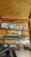 Open wooden case displaying various paint tubes, brushes, and palette inside with paint marks on the lid.