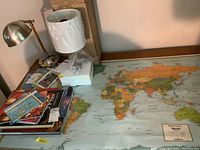 Photo showing a large world map laid flat on a table with colorful country and ocean details, several atlases and books stacked nearby, and two desk lamps—one metallic, one new in box with white shade.