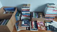 Multiple stacks and boxes of various hardback and paperback books shown on floor and table, titles related to health, cooking, gardening