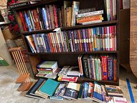 Books stored on wooden bookshelves and stacked on floor showing a variety of fiction and self-help titles, assorted sizes and colors.