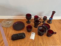 Thirteen pieces of Ruby Red Cape Cod glassware including candlestick, goblets, cups, plates and rectangular dish arranged on the floor with white wall background showing distinctive geometric and hobnail pattern.