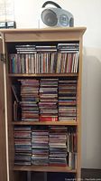Wooden shelf filled with many CDs organized mostly horizontally and vertically, with Emerson CD player on top