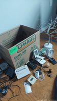 Lot contents arranged on floor with U-Haul small box in background. Visible are wireless camera, cordless phone, desk clock, timers, remote, cables, surge protector, adapter, batteries, and miscellaneous small items.